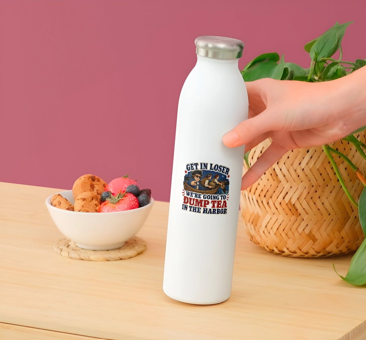 A hand holds a white insulated water bottle with a silver cap on a wooden table. The bottle has a printed graphic with a snake and the text 'GET IN LOSER WE'RE GOING TO DUMP TEA IN THE HARBOR'.