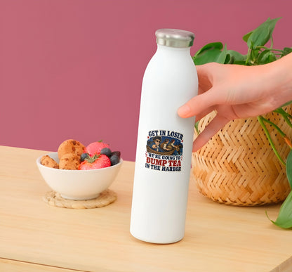 A hand holds a white insulated water bottle with a silver cap on a wooden table. The bottle has a printed graphic with a snake and the text 'GET IN LOSER WE'RE GOING TO DUMP TEA IN THE HARBOR'.
