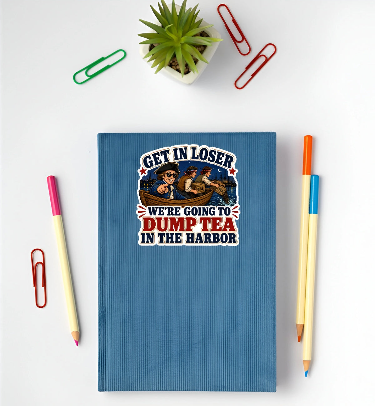A blue textured notebook with a vintage-style cartoon graphic and the text 'GET IN LOSER WE'RE GOING TO DUMP TEA IN THE HARBOR' is shown in a flat-lay arrangement on a white background, surrounded by colorful paperclips, pencils, and a small potted succulent.