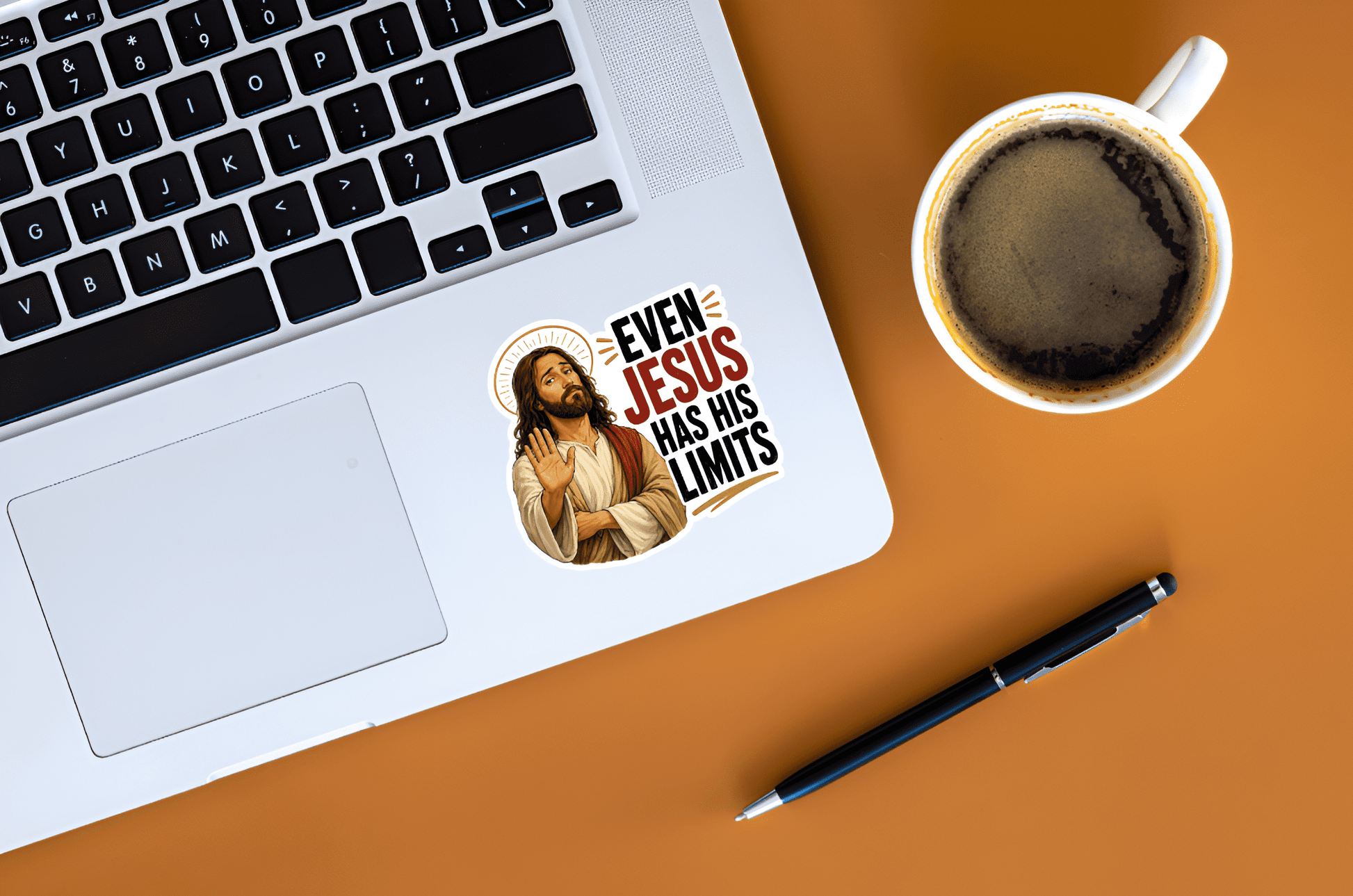 Laptop with a sticker of a illustration of Jesus with a thin gold halo, wearing cream and red robes, holding up one hand in a stopping gesture with a tired expression next to bold stacked text reading "EVEN JESUS HAS HIS LIMITS".and a cup of coffee on a brown surface