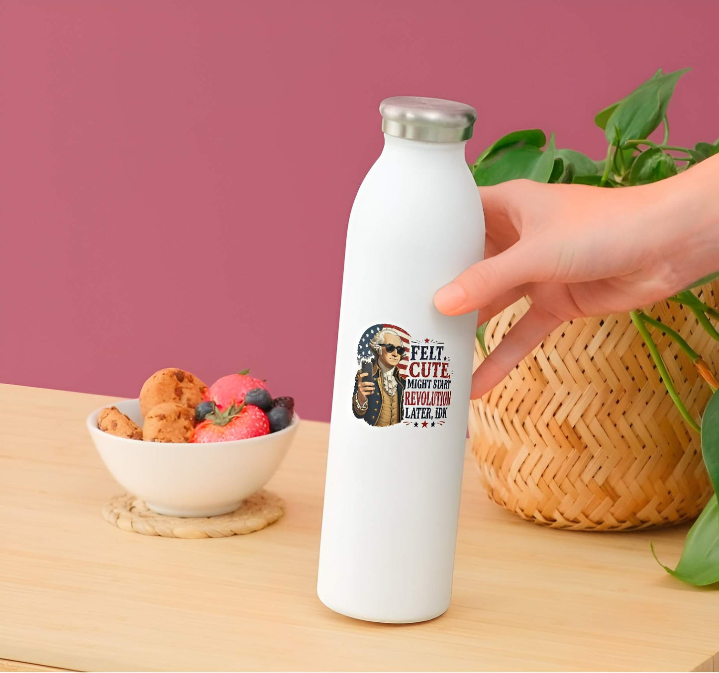 White water bottle with graphic design held by a hand on a wooden table with a pink wall and bowl of snacks in the background.