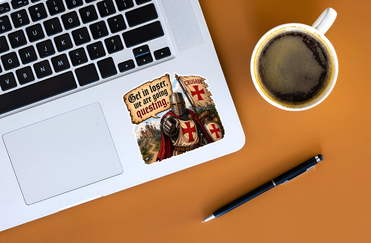 A square sticker with a medieval knight and Templar cross design is shown on the palm rest of a silver laptop. The scene is styled as a flat-lay on an orange surface, accompanied by a cup of coffee and a black pen.