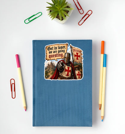 A blue ribbed notebook with a medieval crusader graphic sticker, styled in a flat-lay with colored pencils, paper clips, and a small succulent on a white background.