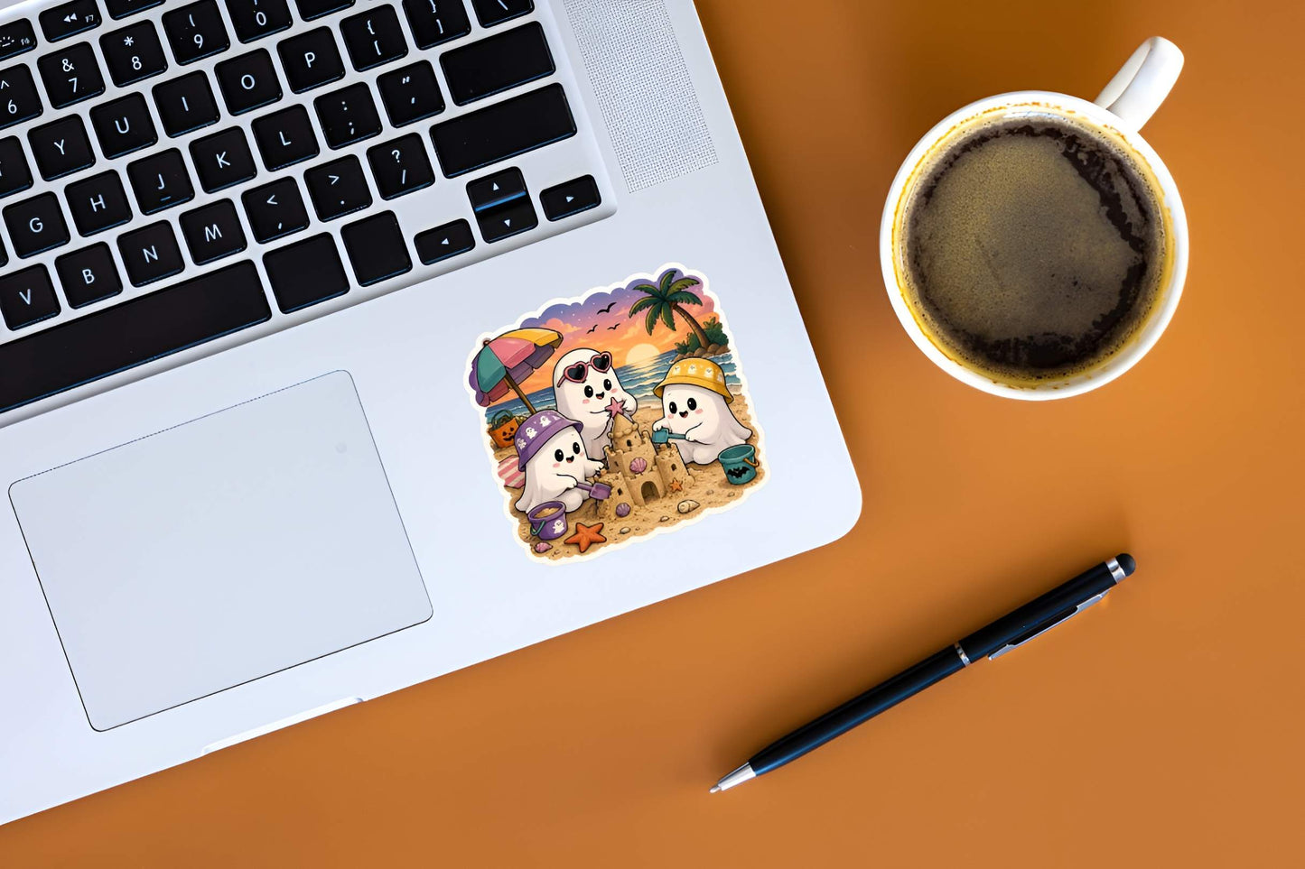 Laptop with a  cute Summerween ghosts sticker, spooky Halloween beach sandcastle vinyl decal, tropical bucket hat ghosts, pink heart sunglasses, palm tree sunset graphic, a cup of coffee, and a pen on a brown surface.