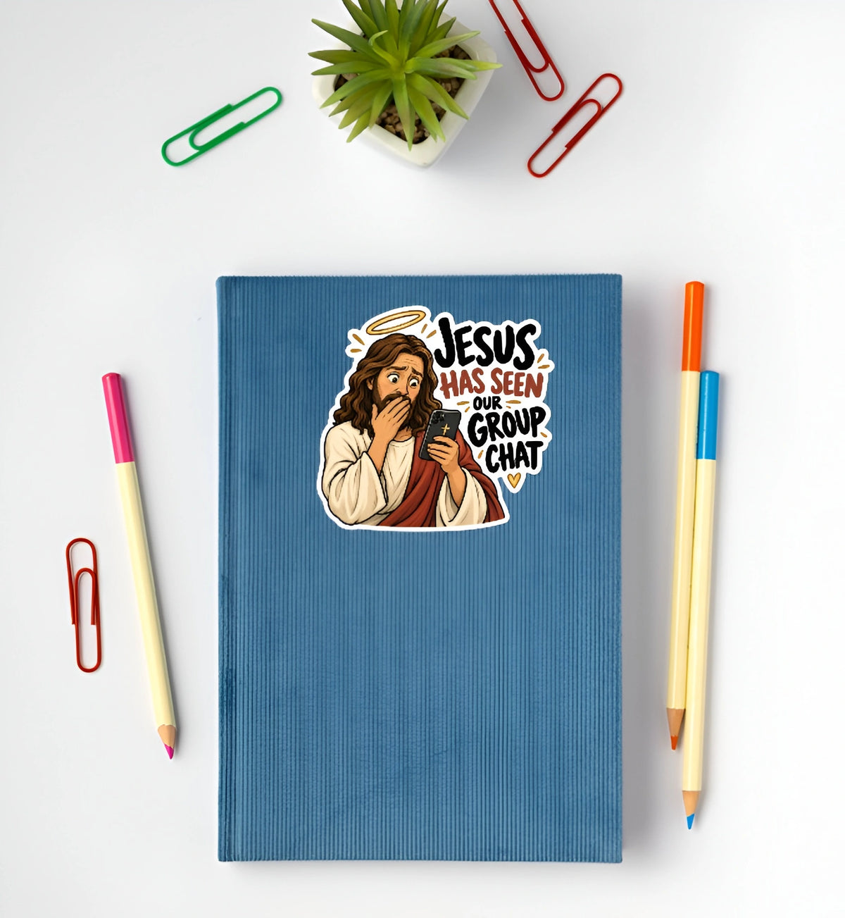 A blue ribbed notebook with a cartoon sticker of Jesus covering his mouth and the text 'JESUS HAS SEEN OUR GROUP CHAT' lies on a white desk surrounded by office props including paper clips, pencils, and a small potted plant.
