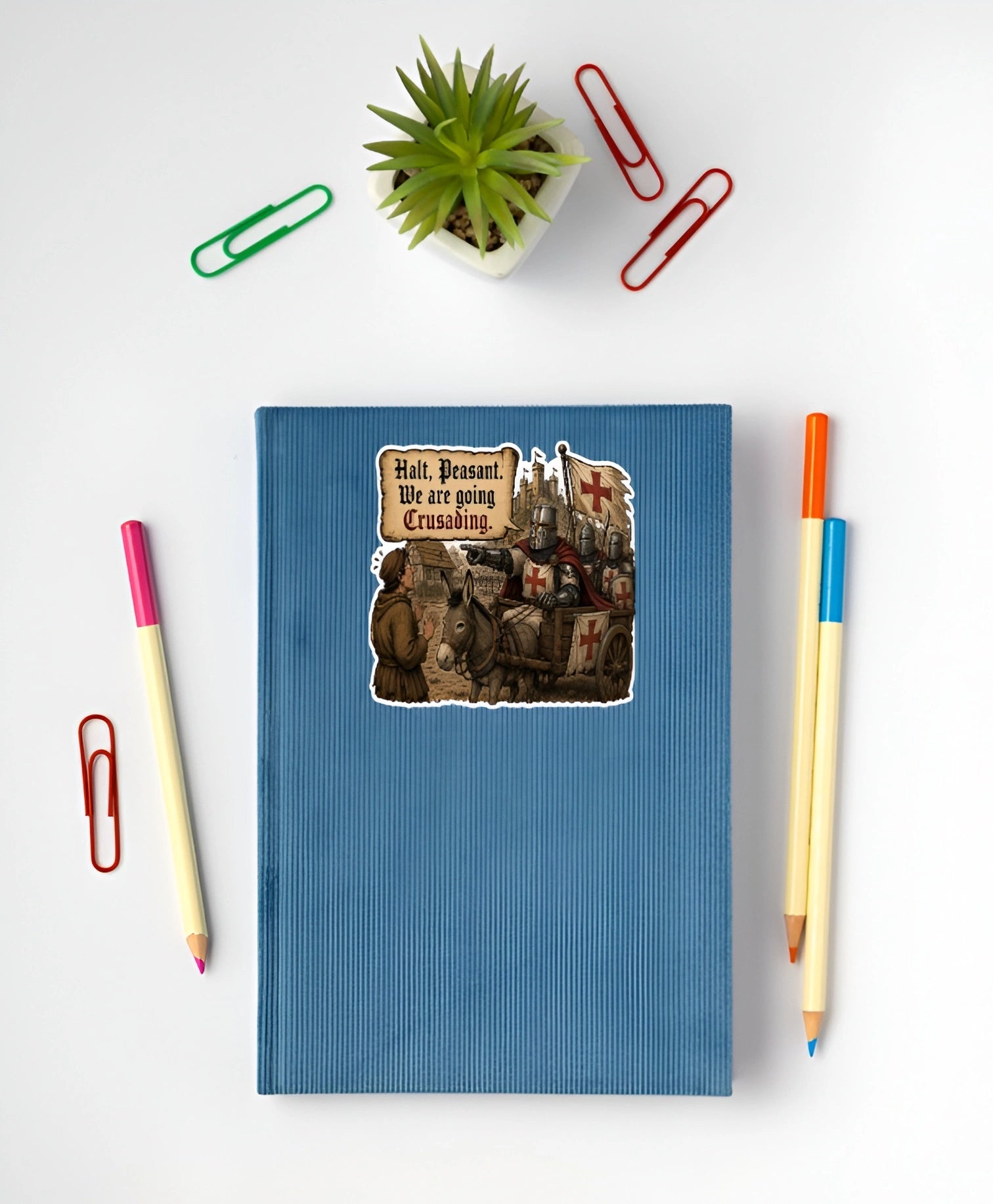 Funny medieval crusading sticker with knights and donkey cart displayed on a blue notebook