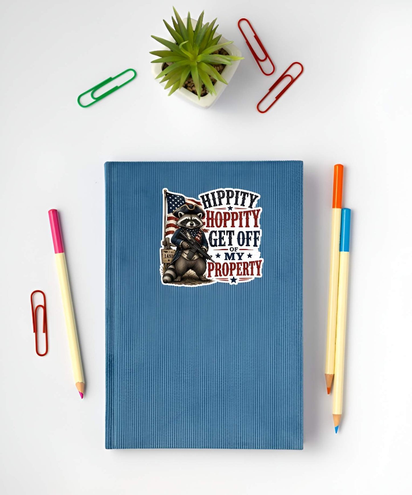 Blue notebook with a graphic design featuring a raccoon and text, surrounded by stationery items on a white background.
