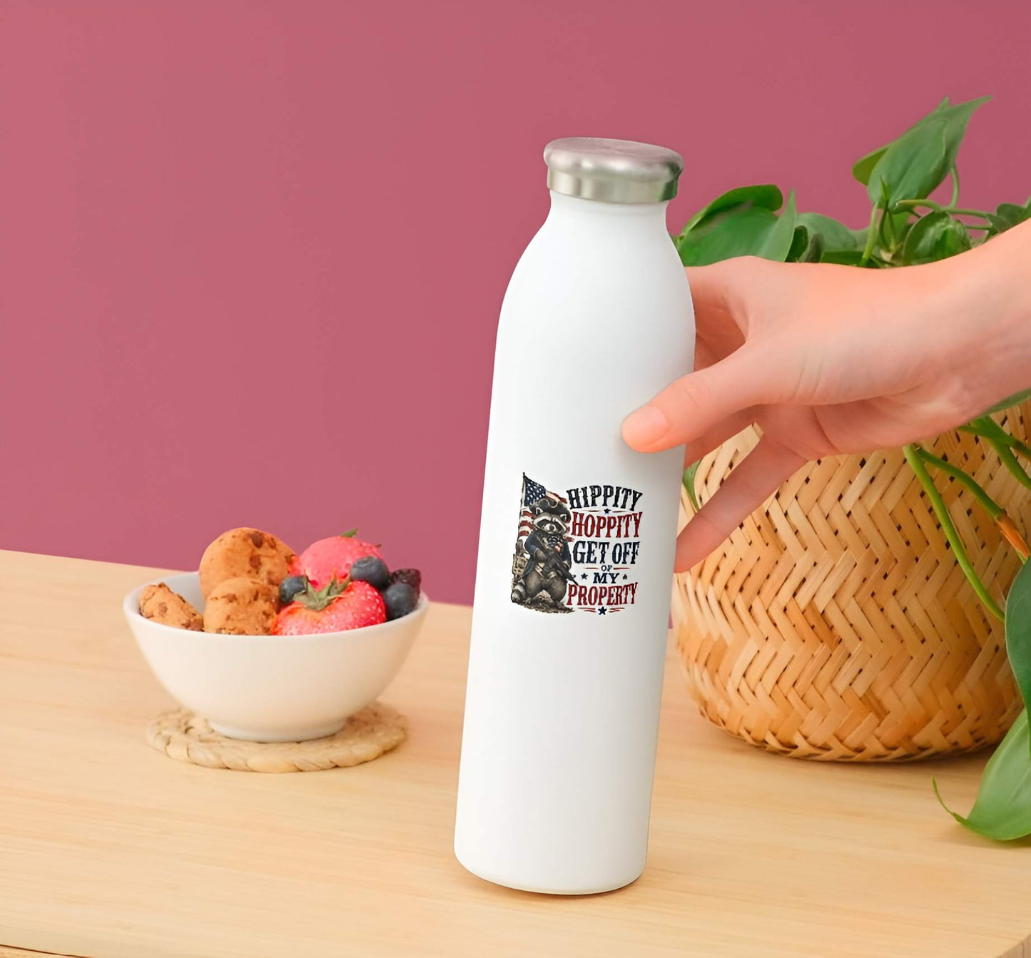 White water bottle with a printed design held by a hand on a wooden table with a bowl of snacks and a plant in the background.