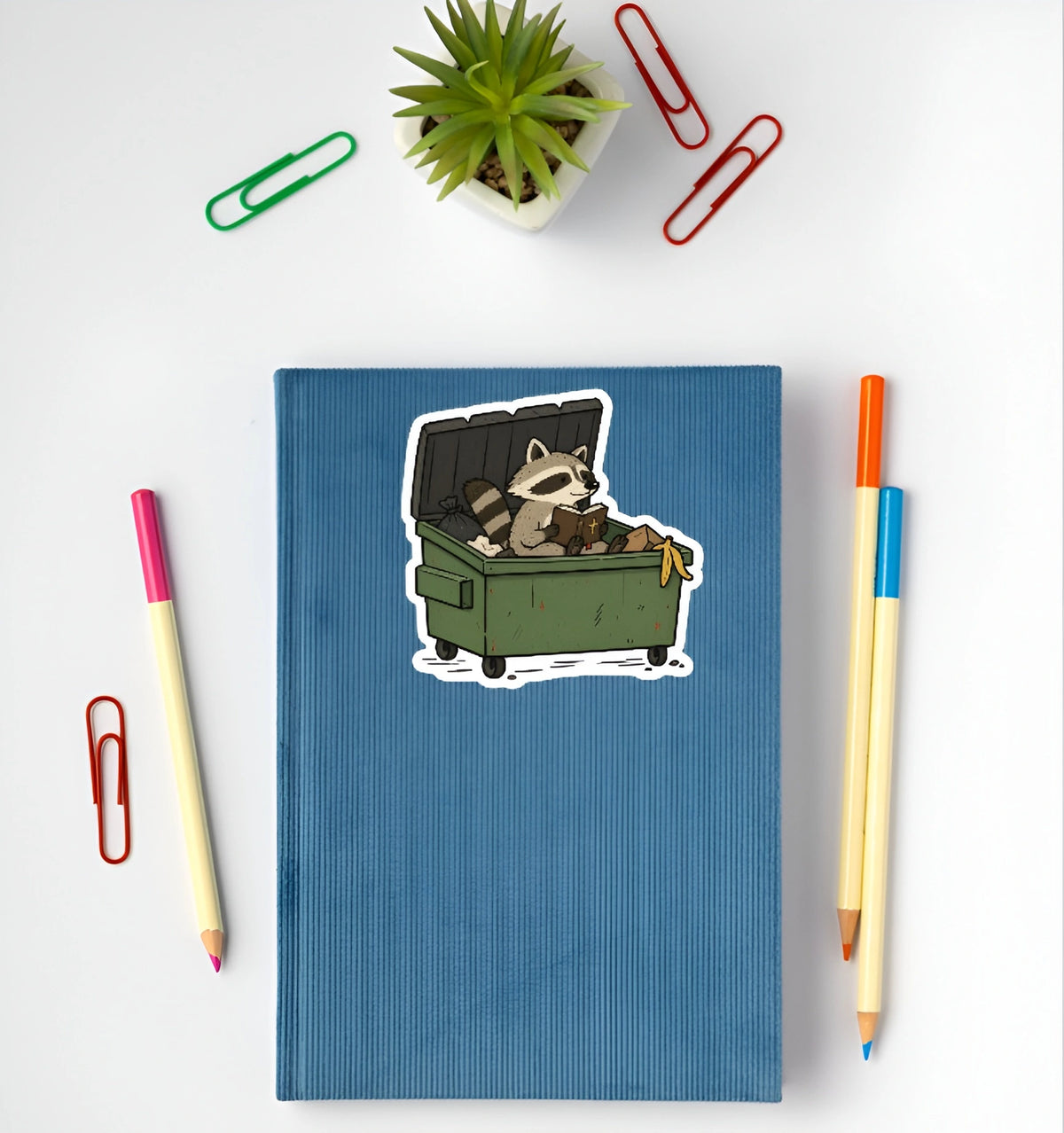 Funny raccoon Bible sticker featuring a raccoon sitting in a green dumpster displayed on a blue notebook