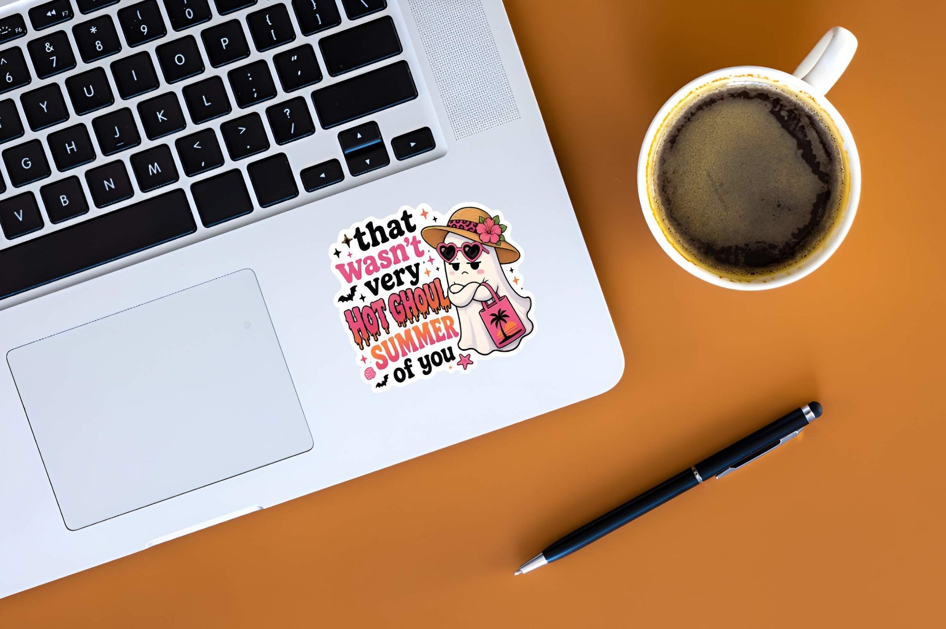 Laptop with a Funny Hot Ghoul Summer ghost sticker, cute Halloween beach vinyl decal, tropical ghost in straw hat and pink heart sunglasses with sunset beach tote bag graphic. on a desk next to a cup of coffee and pen