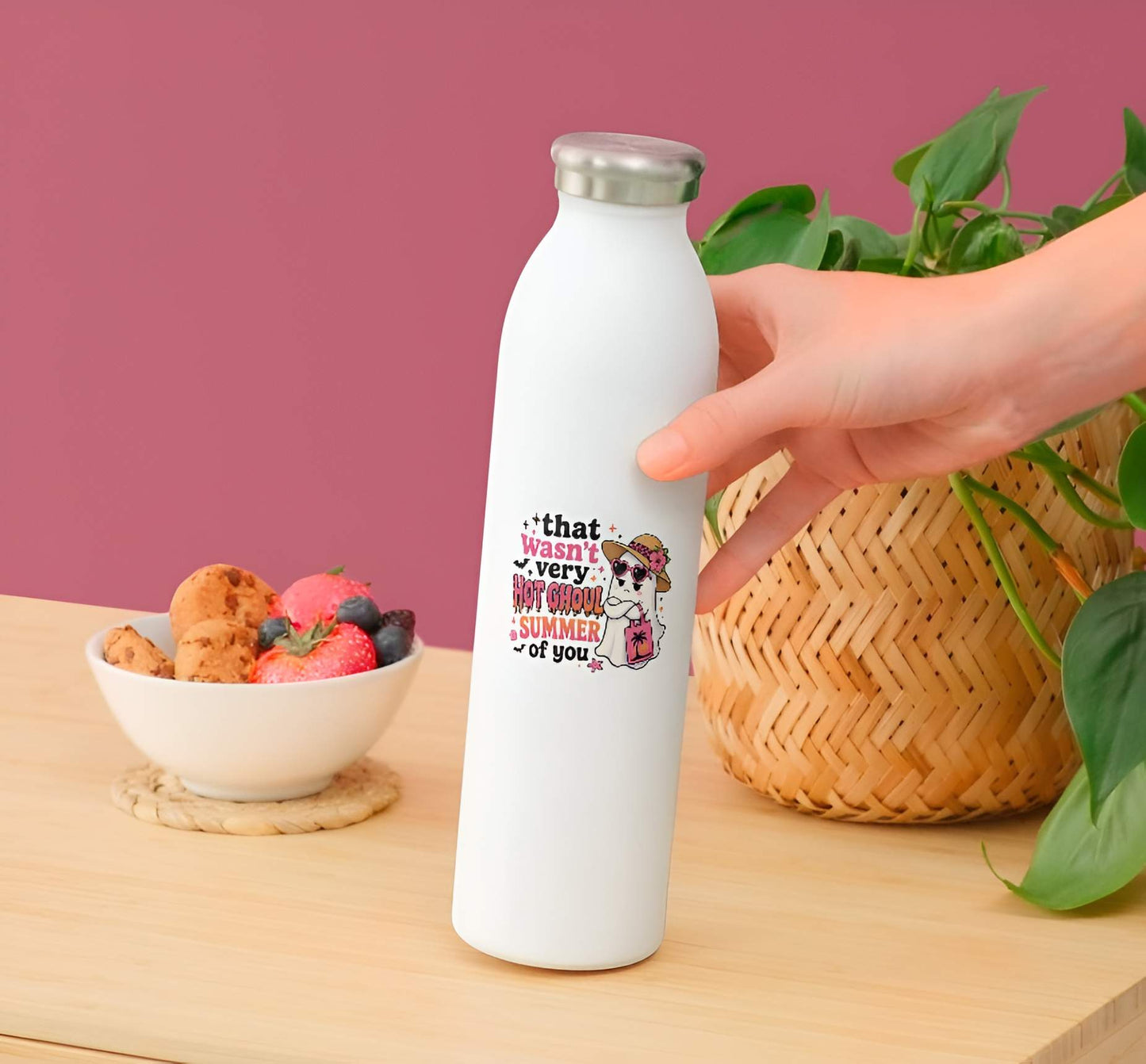 White water bottle withFunny Hot Ghoul Summer ghost sticker, cute Halloween beach vinyl decal, tropical ghost in straw hat and pink heart sunglasses with sunset beach tote bag graphic.  held by a hand on a wooden surface with a bowl of fruit and plant in the background.