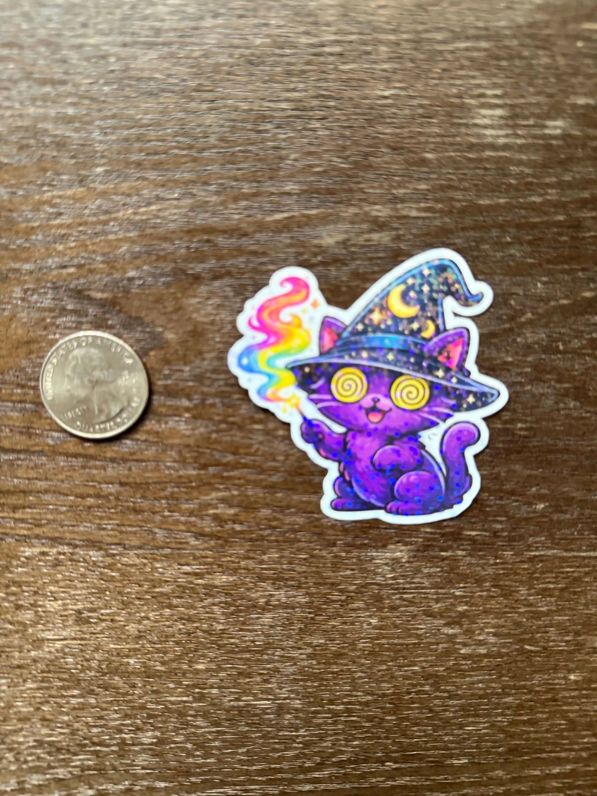 Colorful cat sticker with a witch hat on a wooden surface next to a coin for scale.