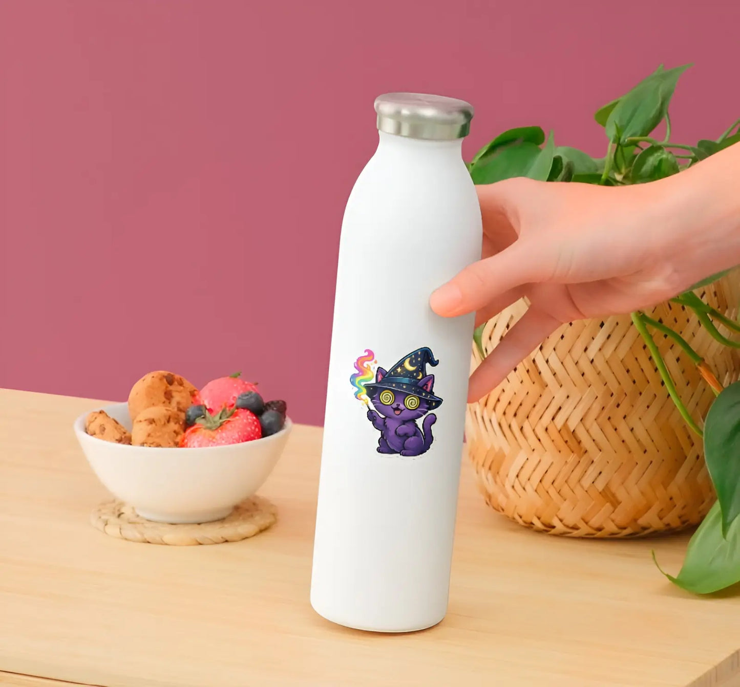 White water bottle with a colorful cat design held by a hand on a wooden table with a bowl of snacks and plant in the background.