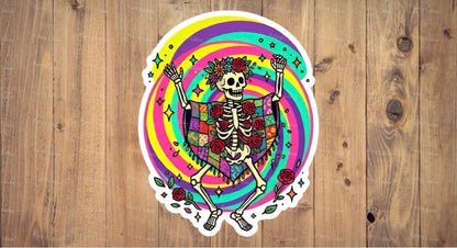 A flat, isolated holographic vinyl sticker based on image.png featuring a joyful cartoon skeleton dancing with open arms against a swirling rainbow spiral backdrop, wearing a colorful patchwork poncho and a red rose flower crown.