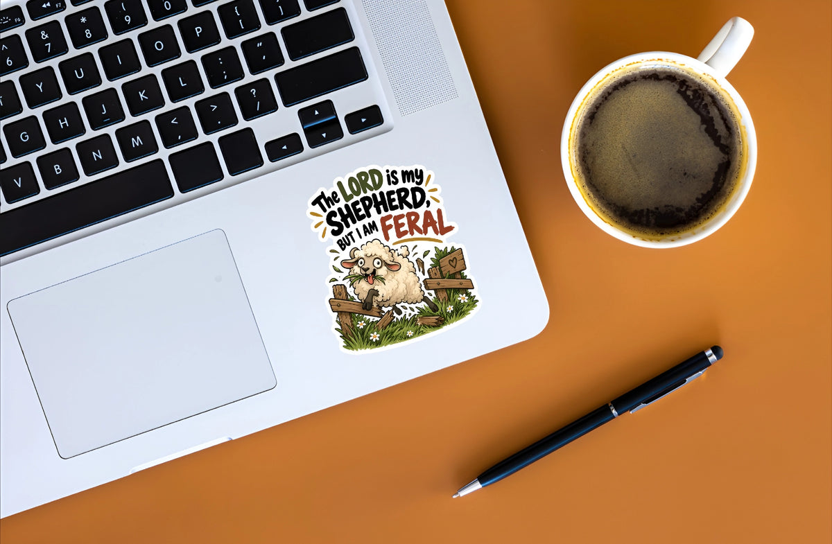 A die-cut vinyl sticker with the text 'THE LORD IS MY SHEPHERD BUT I'M FERAL' and a cartoon sheep sitting in grass, shown on the palm rest of a silver laptop. The laptop is part of a flat-lay arrangement on a solid orange surface, alongside a white coffee mug and a black pen.