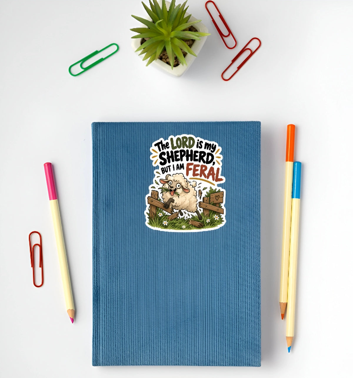 A blue ribbed hardcover notebook with a cartoon sheep sticker on the front, styled in a flat lay with pens, paperclips, and a small potted succulent on a white background.