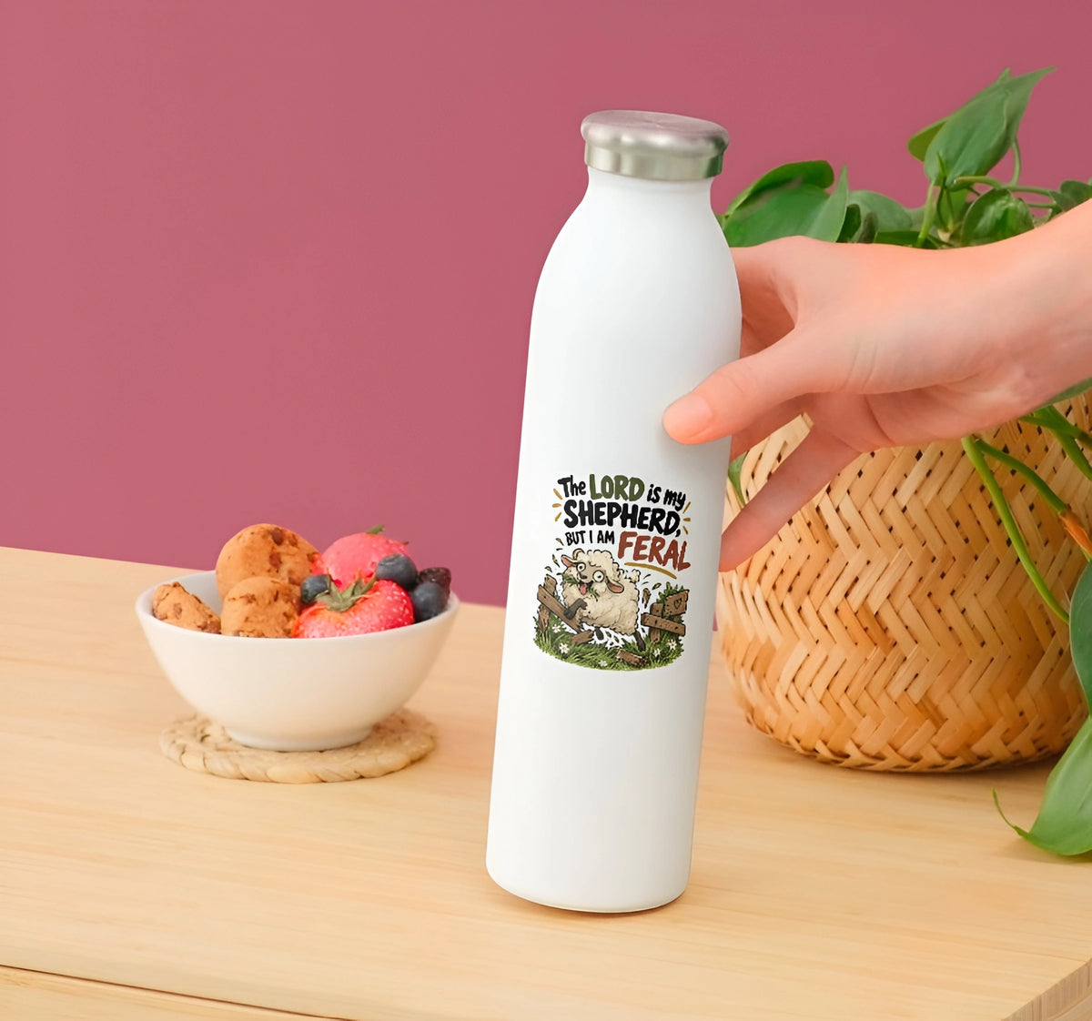 A hand holds a white insulated water bottle with a silver cap on a wooden table. The bottle features a graphic of a sheep and the text 'THE LORD IS MY SHEEPHERD: I AM FERAL'.