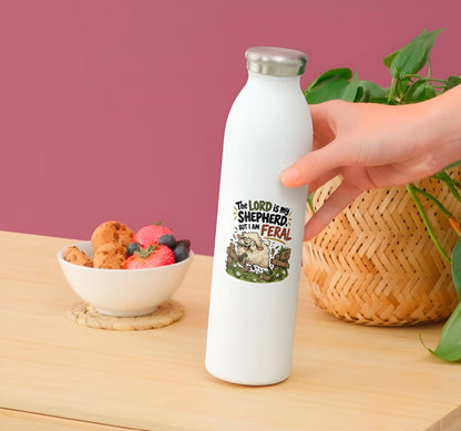 A hand holds a white insulated water bottle with a silver cap on a wooden table. The bottle features a graphic of a sheep and the text 'THE LORD IS MY SHEEPHERD: I AM FERAL'.