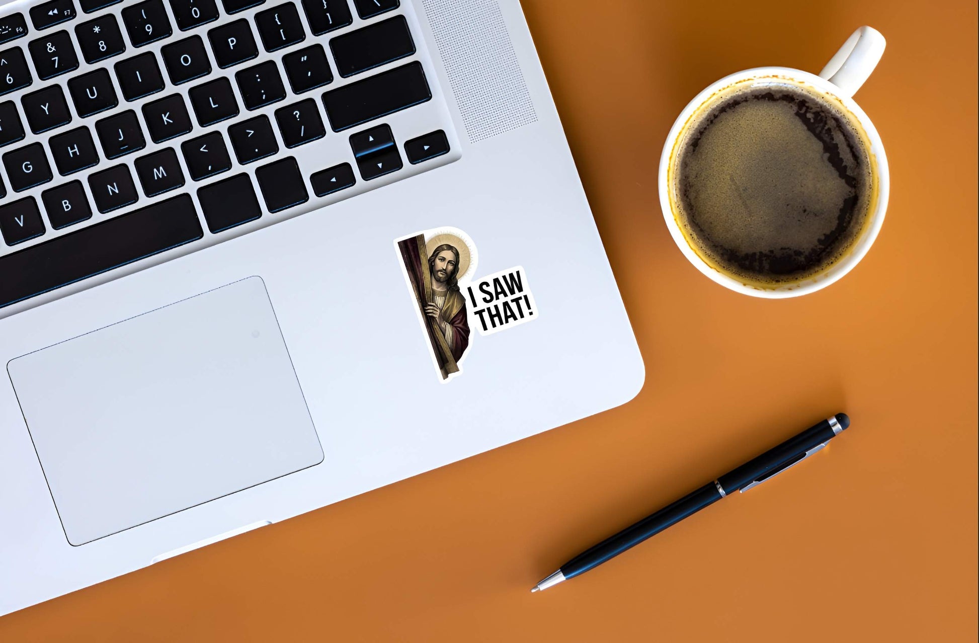 Laptop with a sticker of a Jesus Christ with 'I Saw That!' text, coffee cup, and pen on an orange surface