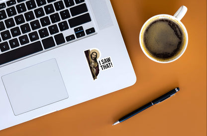 Laptop with a sticker of a Jesus Christ with 'I Saw That!' text, coffee cup, and pen on an orange surface