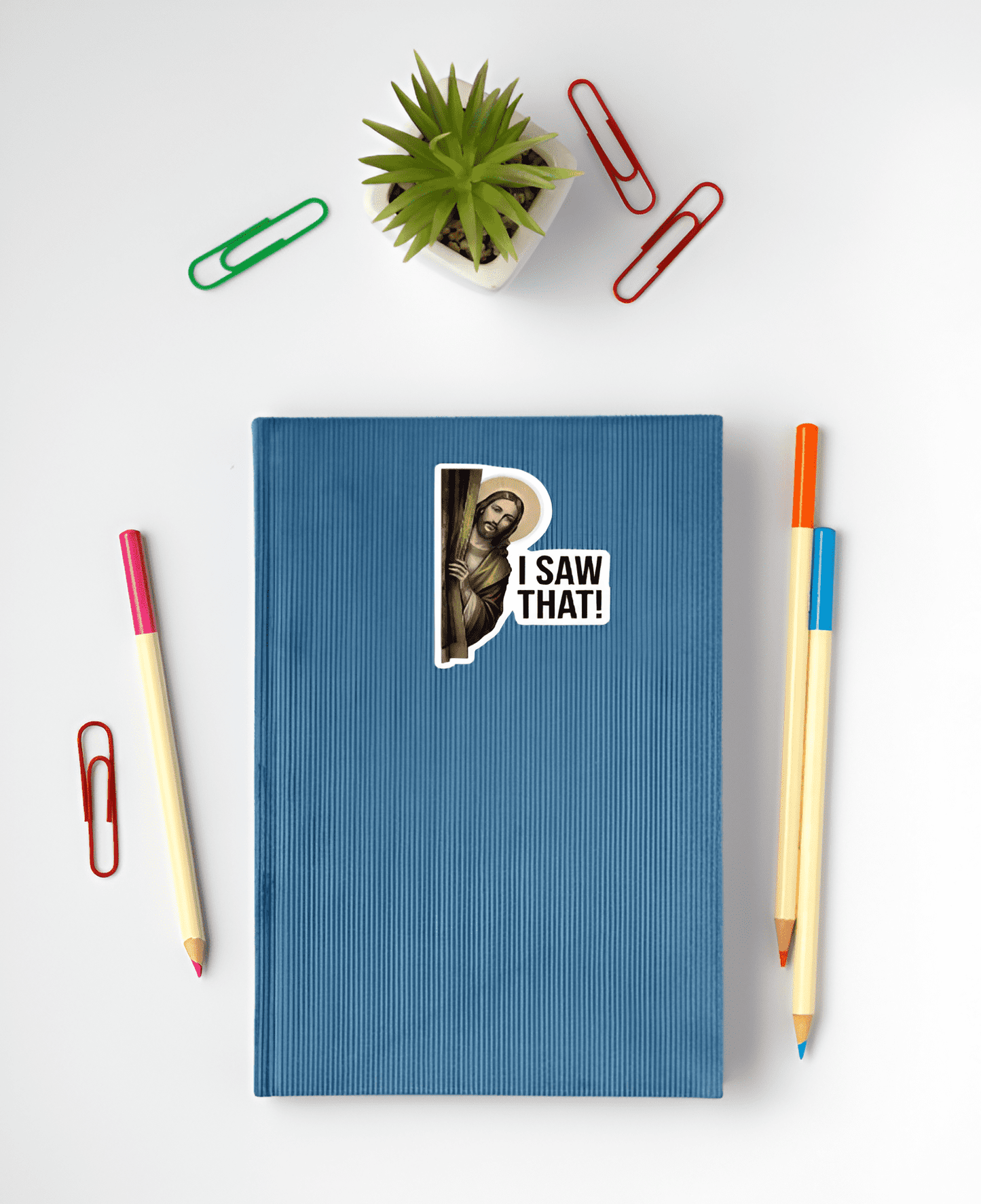 Blue notebook with a sticker of a Jesus Christ with 'I Saw That!' text on a white background with stationery items.