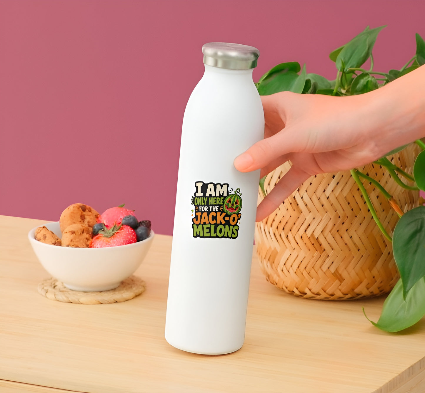 White water bottle with a cFunny Summerween watermelon sticker by a hand on a wooden surface with a bowl of snacks and plant in the background.