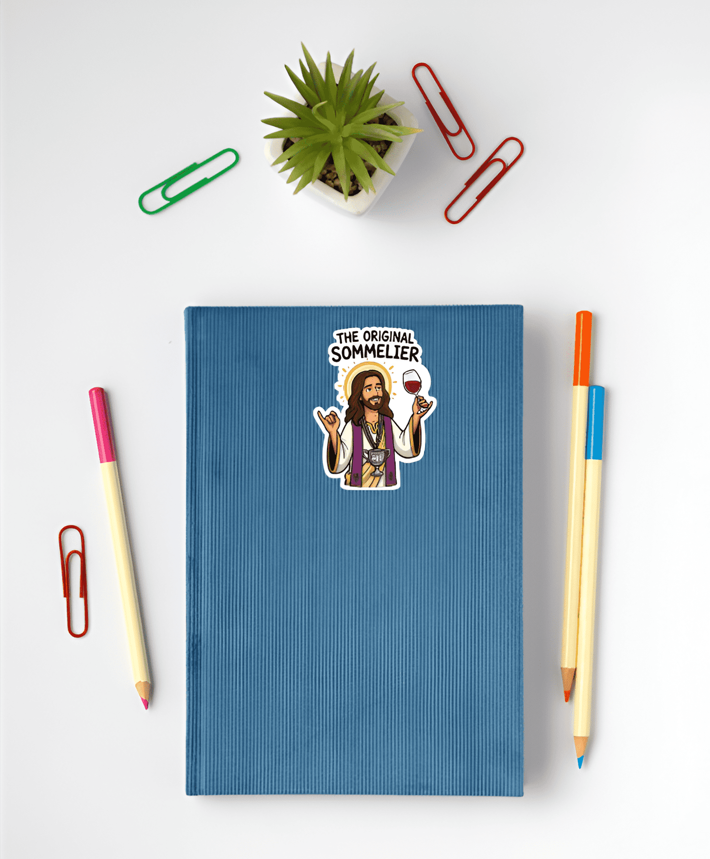 Blue notebook with a sticker of a Jesus labeled 'The Original Sommelier' on a white surface with stationery items.