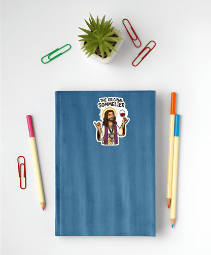 Blue notebook with a sticker of a Jesus labeled 'The Original Sommelier' on a white surface with stationery items.