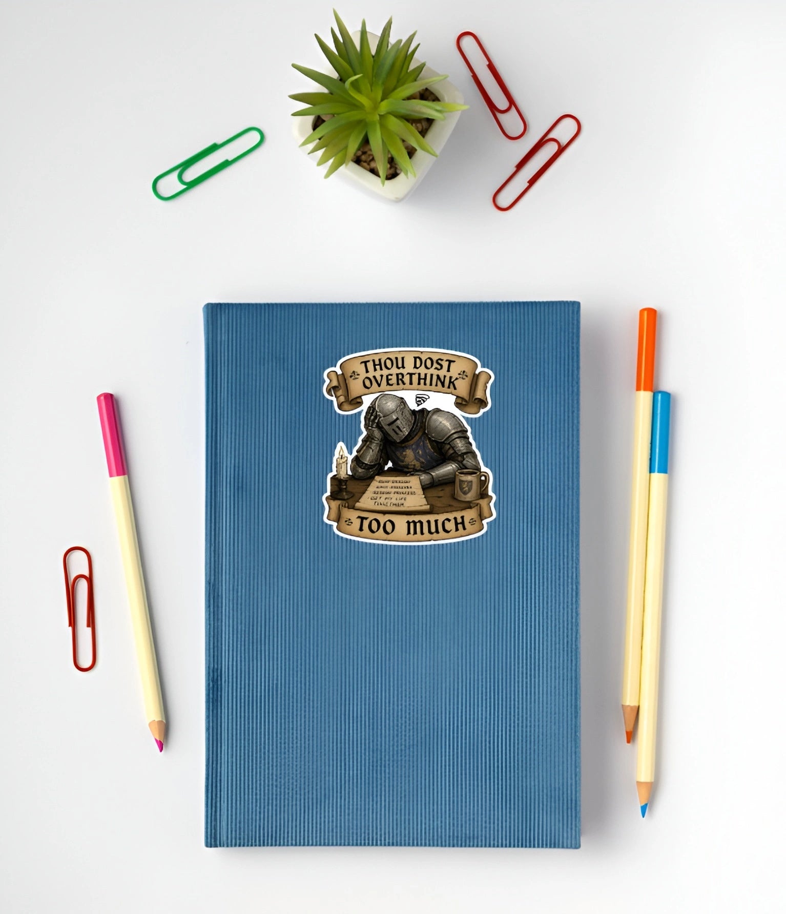 A blue ribbed notebook with a humorous sticker that reads 'THOU DOST OVERTHINK TOO MUCH' featuring an illustration of a knight in armor. The notebook is styled in a flat-lay arrangement on a white background with colorful paperclips, a small potted succulent, and two pencils.
