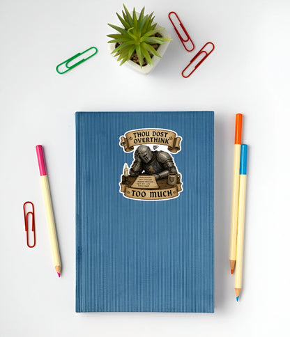 A blue ribbed notebook with a humorous sticker that reads 'THOU DOST OVERTHINK TOO MUCH' featuring an illustration of a knight in armor. The notebook is styled in a flat-lay arrangement on a white background with colorful paperclips, a small potted succulent, and two pencils.