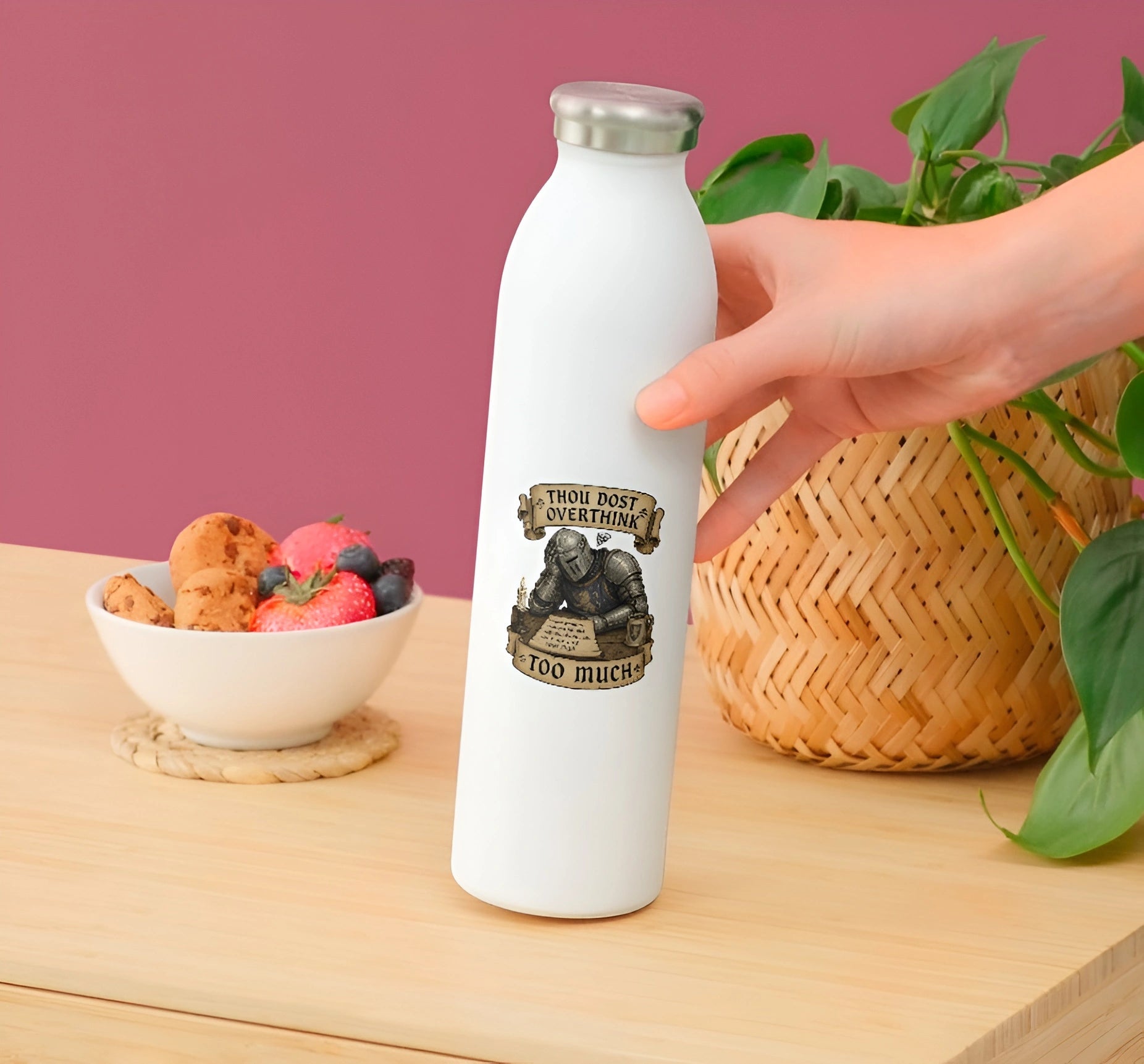 A hand holding a white insulated water bottle with a silver cap, featuring a graphic of a knight and the text 'THOU SHIP WEARABLE TOO MUCH'. The bottle is set on a light wood table with a pink background, flanked by a bowl of fruit and a potted plant.