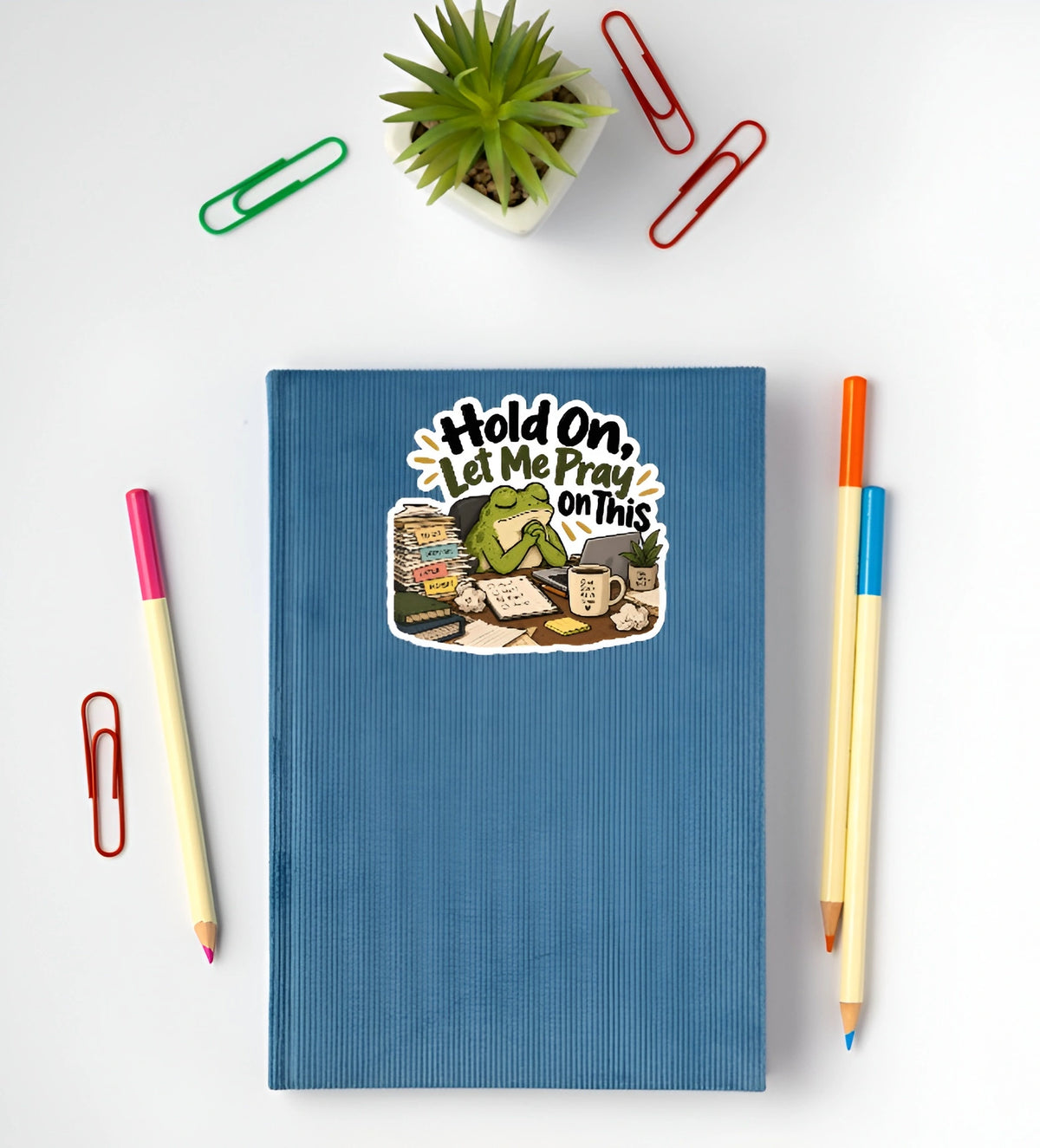A blue ribbed notebook with a cartoon sticker that reads 'Hold On, Let Me Pray On This' is shown in a flat-lay arrangement with colored pencils, paperclips, and a small potted succulent on a white background.
