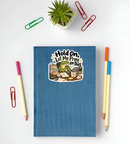 A blue ribbed notebook with a cartoon sticker that reads 'Hold On, Let Me Pray On This' is shown in a flat-lay arrangement with colored pencils, paperclips, and a small potted succulent on a white background.