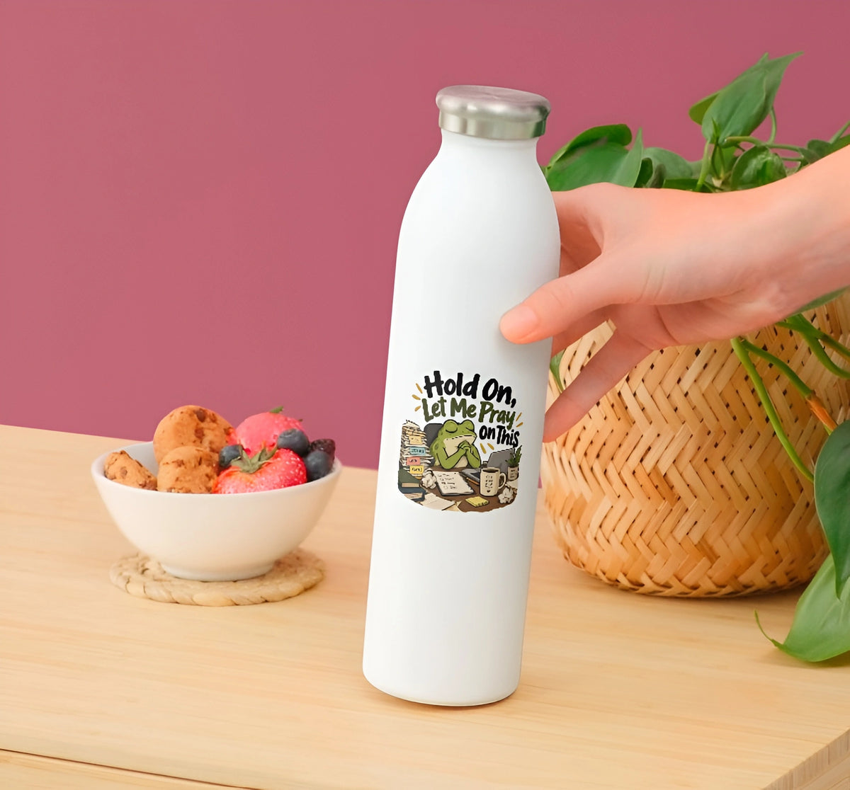 A white insulated water bottle with a silver cap stands on a wooden table, with a hand holding the bottle. The bottle has a graphic print that reads 'Hold On Let Me Pray On This' with a Bible illustration.