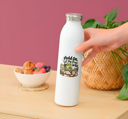 A white insulated water bottle with a silver cap stands on a wooden table, with a hand holding the bottle. The bottle has a graphic print that reads 'Hold On Let Me Pray On This' with a Bible illustration.