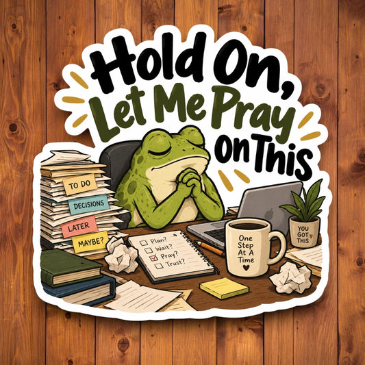 A die-cut sticker featuring a green frog praying at a cluttered desk with the text 'Hold On, Let Me Pray On This' in bold black and olive lettering, shown against a brown wood plank background.