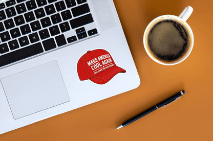Laptop with 'Make America Cool Again' sticker, coffee cup, and pen on a brown surface