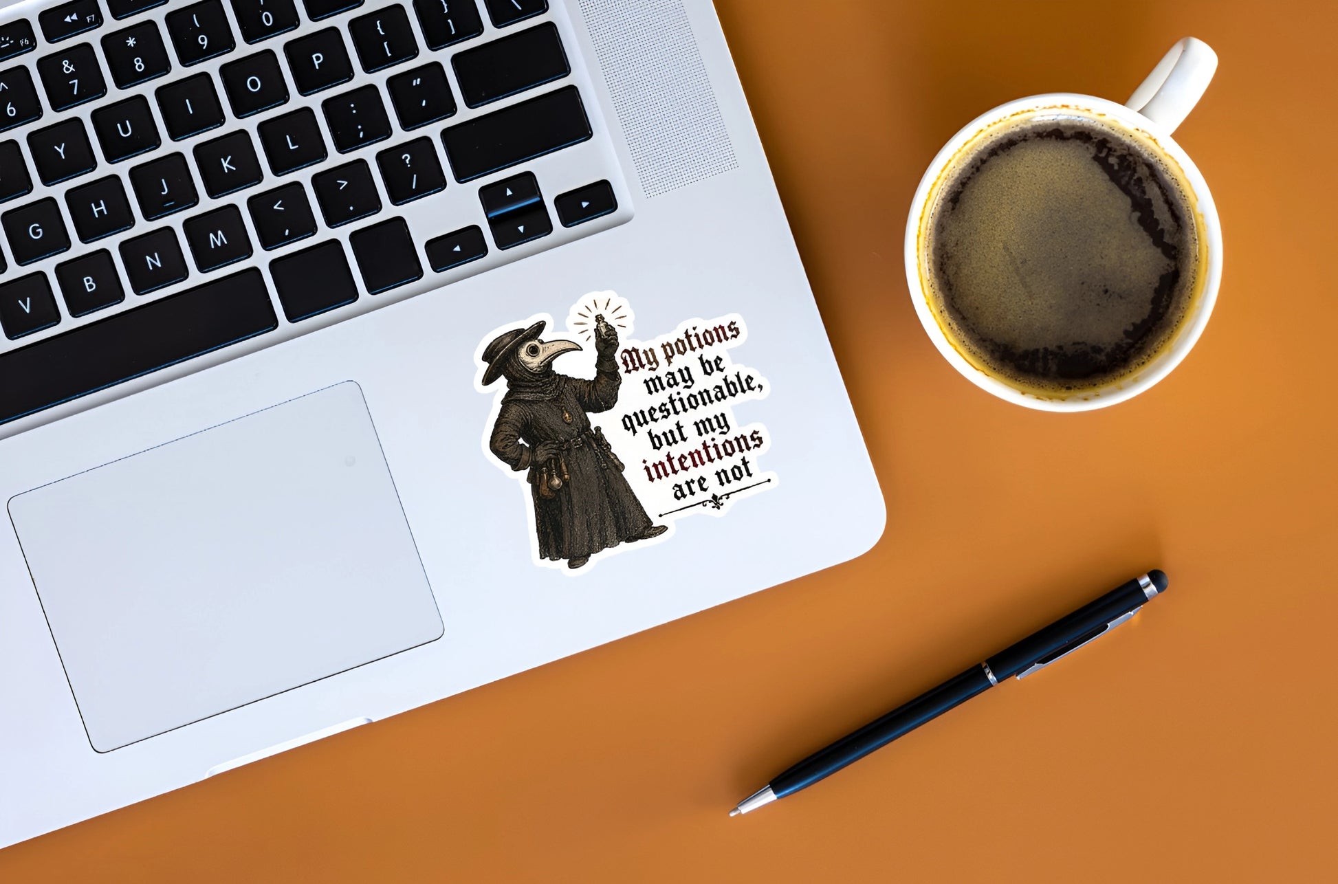 A black and white vintage-style sticker with a witch illustration and the quote 'My potions may be questionable, but my intentions are not' applied to the palm rest of a silver laptop. The laptop is styled on an orange desk alongside a cup of coffee and a black pen.