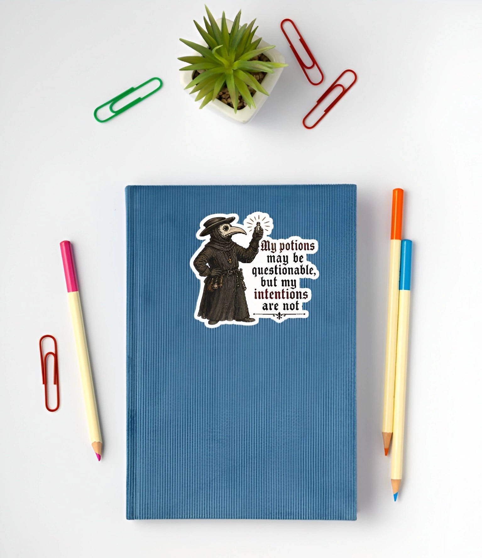 A blue ribbed notebook with a vintage-style cartoon sticker of a plague doctor and the quote 'My potions may be questionable, but my intentions are not', shown in a flat-lay arrangement with colored paperclips, a small potted succulent, and two pencils on a white background.