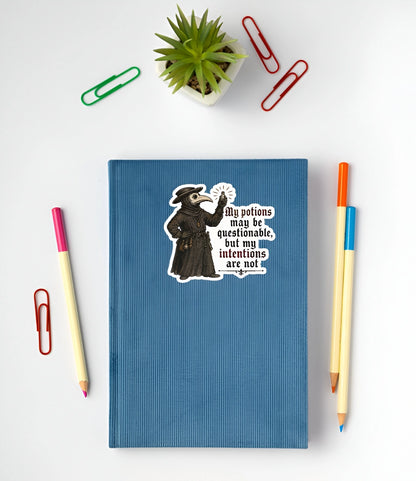 A blue ribbed notebook with a vintage-style cartoon sticker of a plague doctor and the quote 'My potions may be questionable, but my intentions are not', shown in a flat-lay arrangement with colored paperclips, a small potted succulent, and two pencils on a white background.