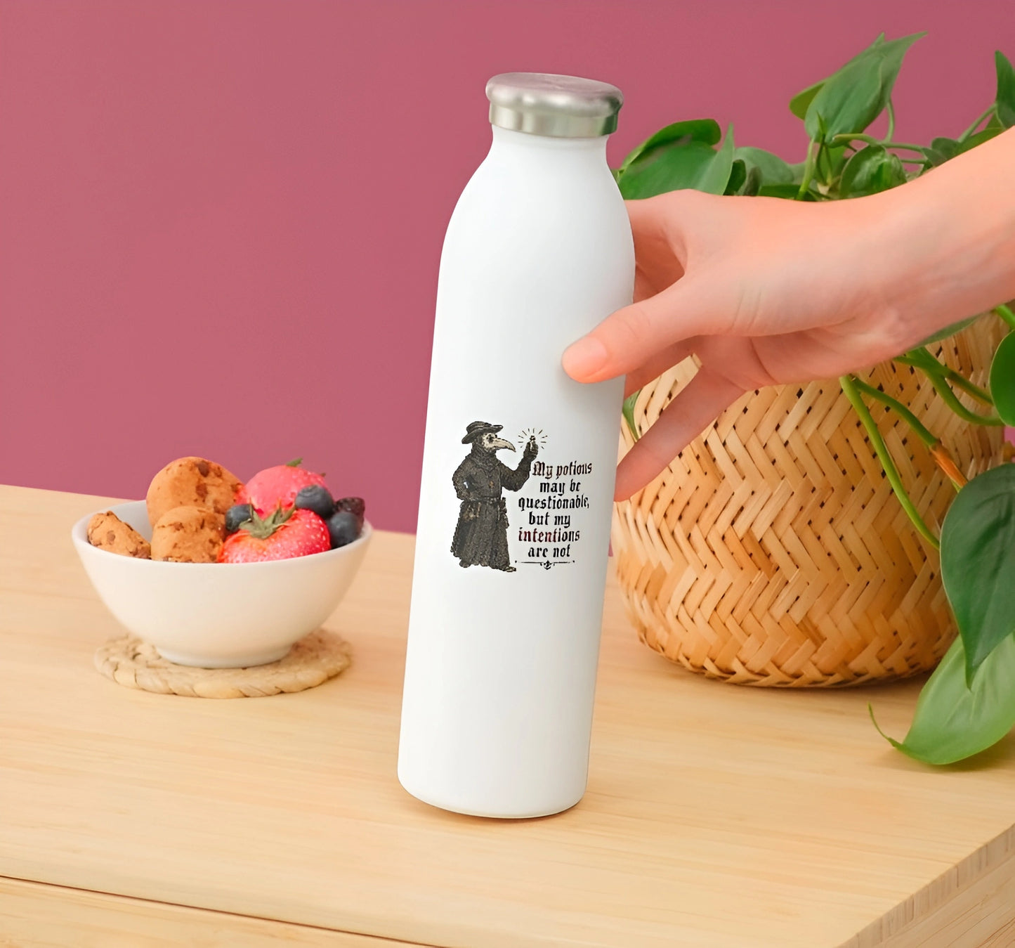 A hand holds a white insulated water bottle with a silver cap on a wooden table. The bottle has a black graphic of a rooster 