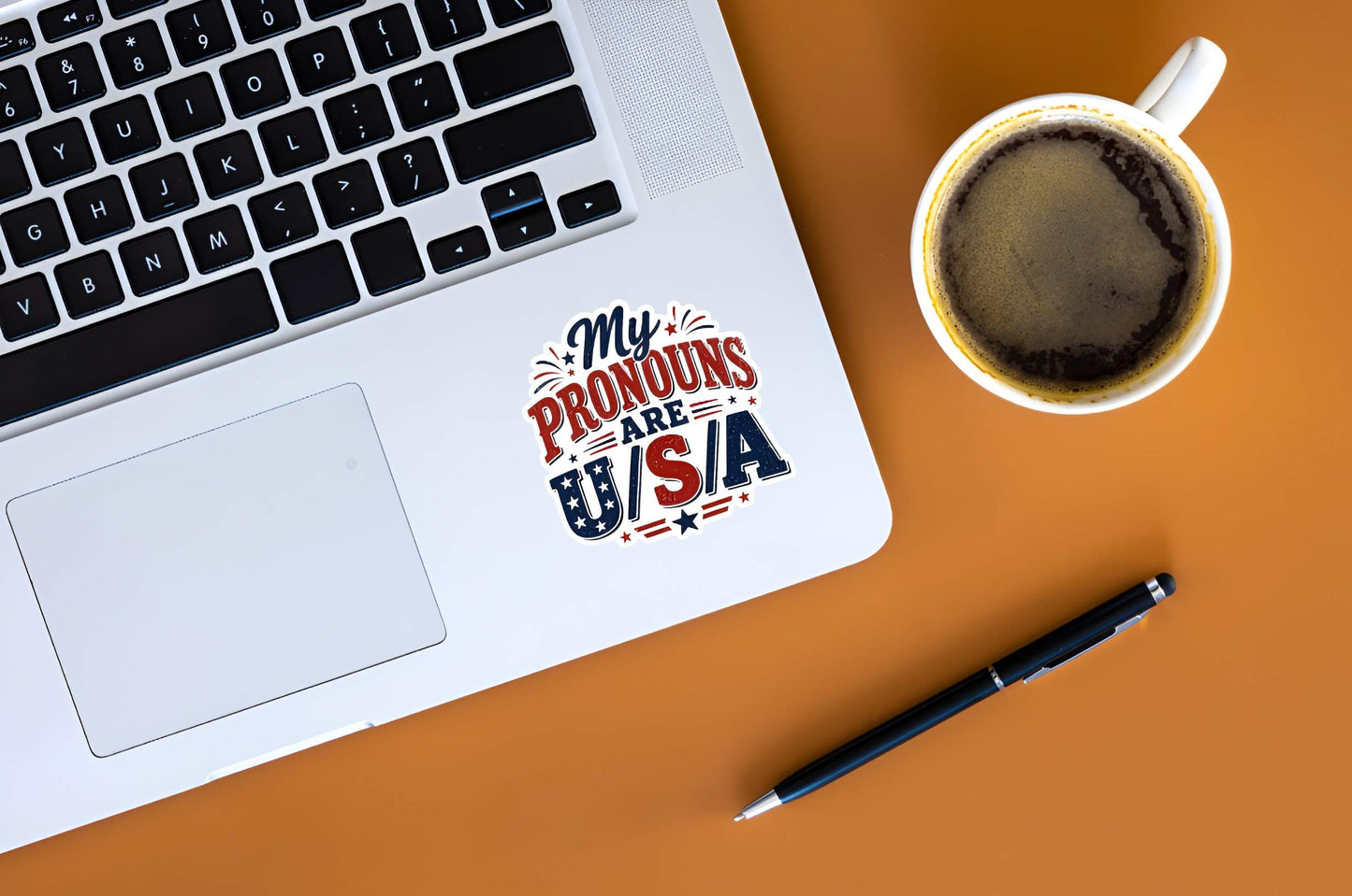 Laptop with a 'My Pronouns Are USA' sticker, cup of coffee, and pen on an orange surface