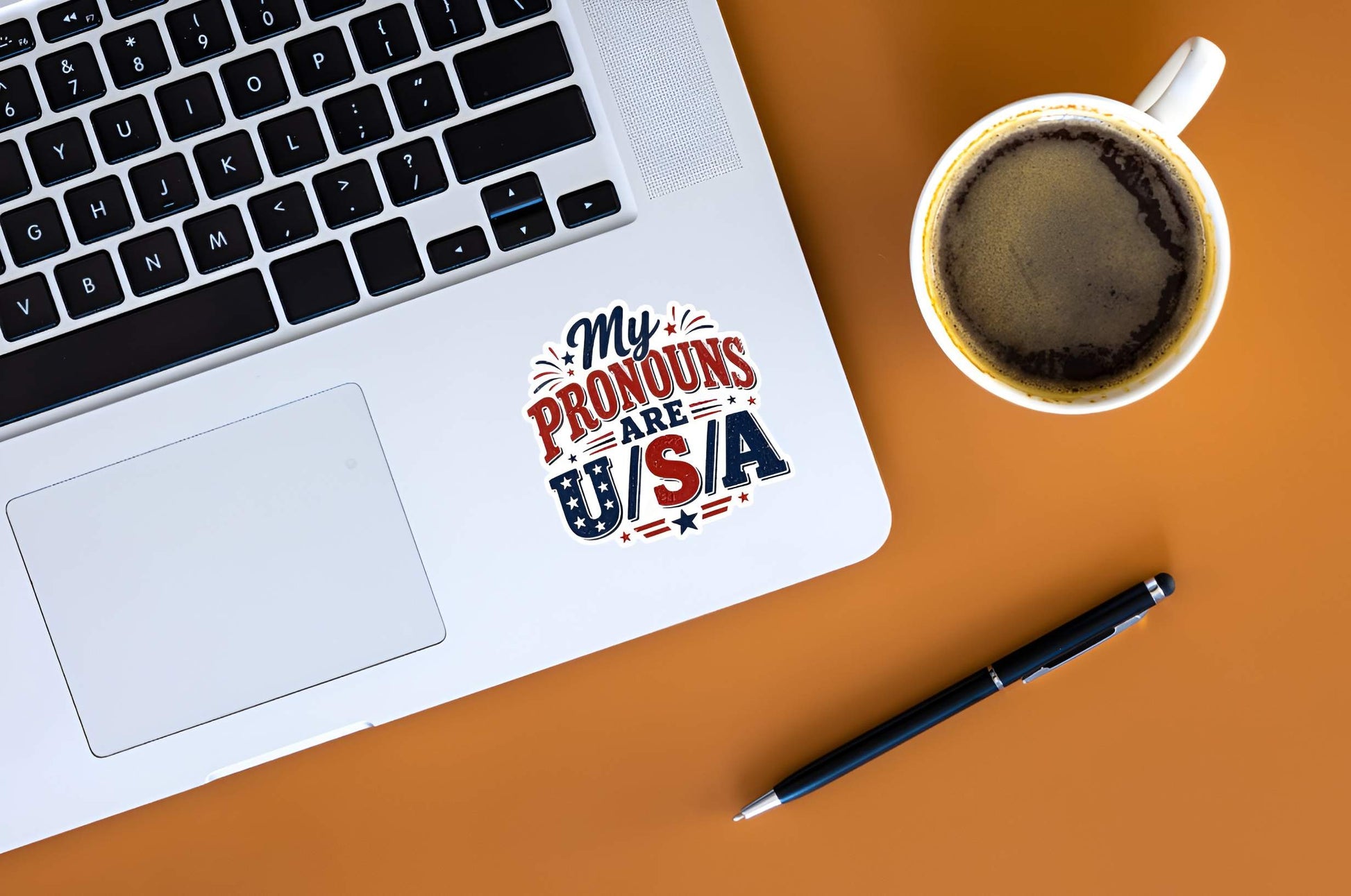 Laptop with a 'My Pronouns Are USA' sticker, cup of coffee, and pen on an orange surface