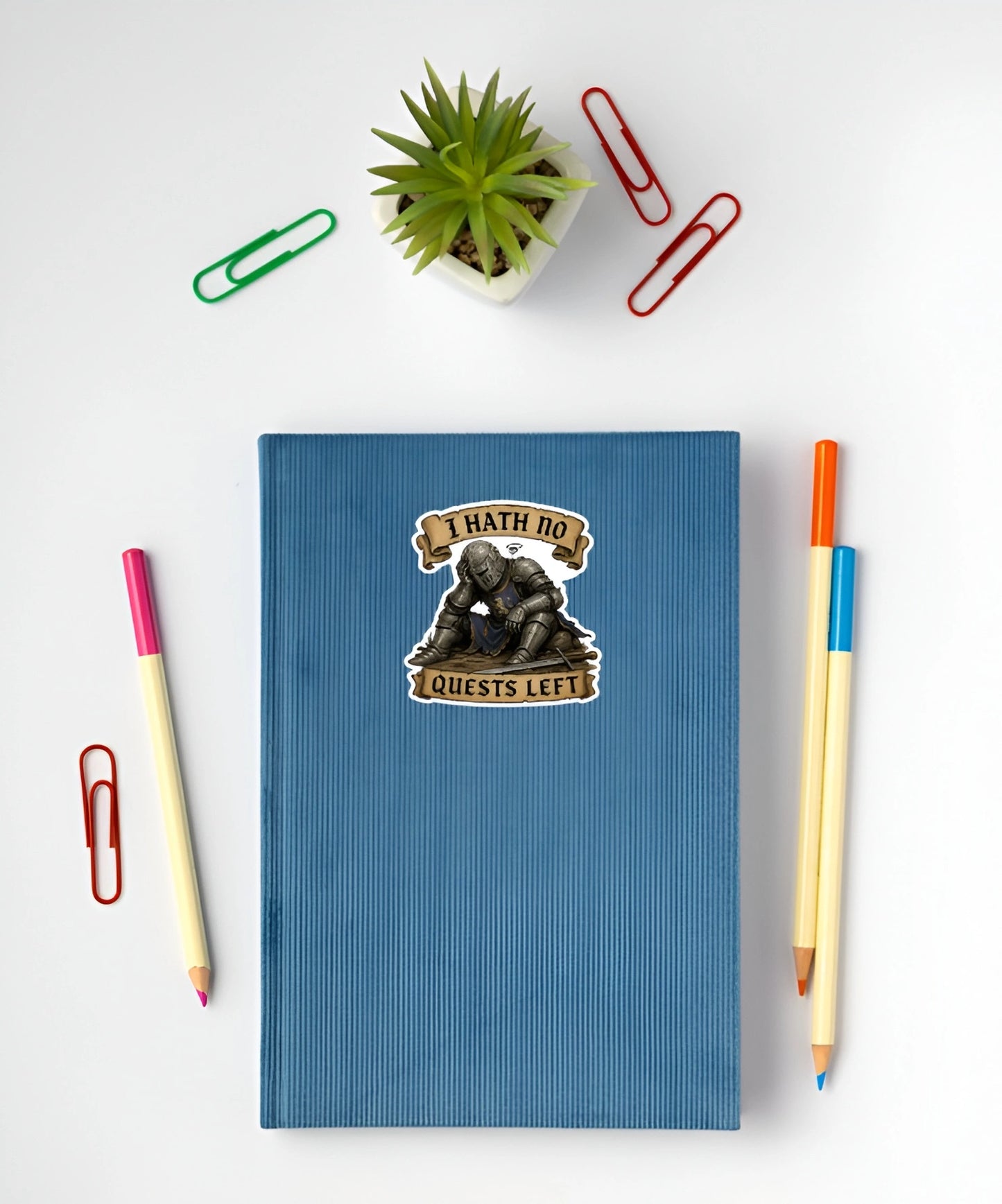 A blue ribbed notebook with a 'I HATH NO QUESTS LEFT' knight sticker, styled in a flat lay with pencils, paper clips, and a small potted plant on a white background.
