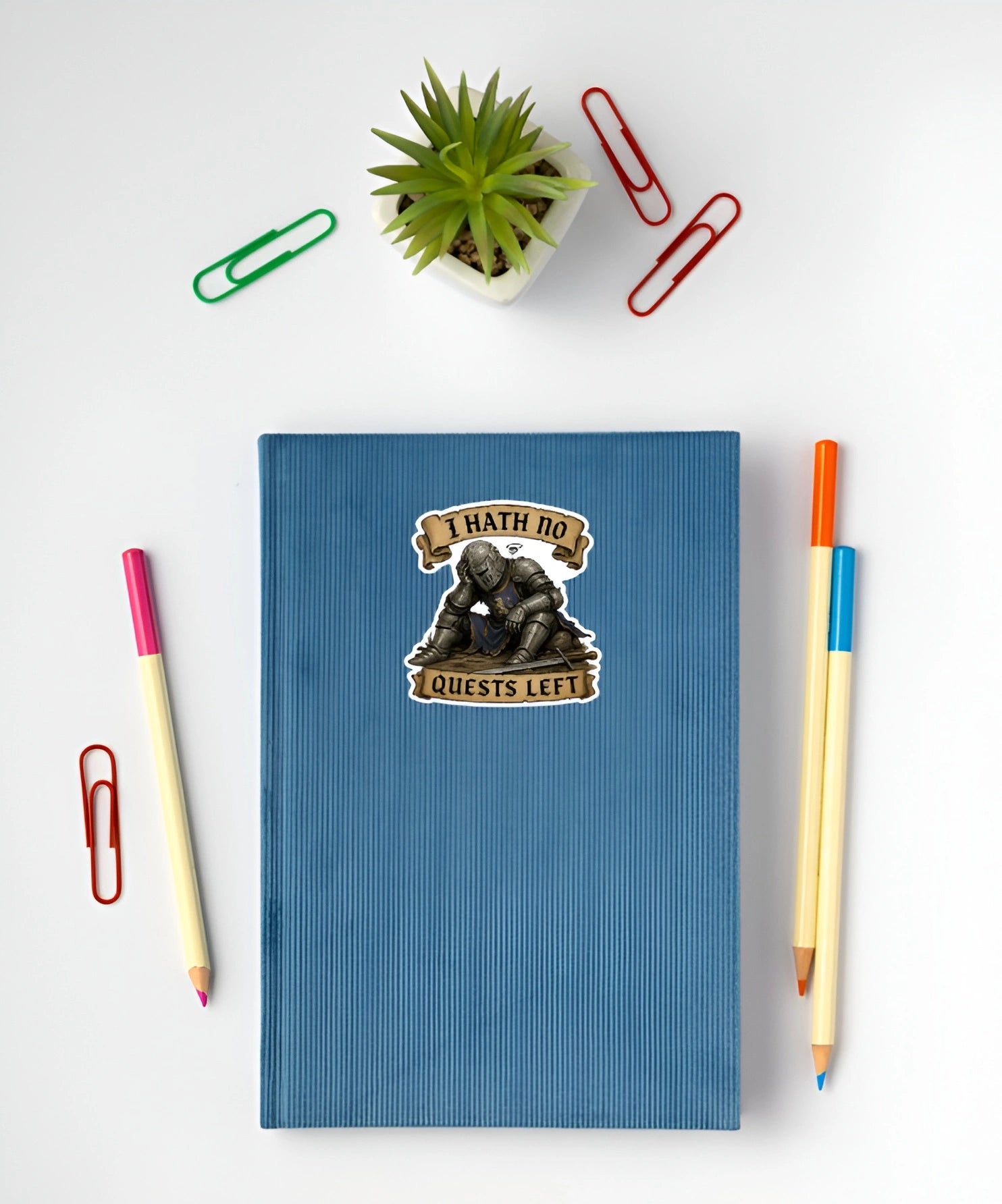 A blue ribbed notebook with a 'I HATH NO QUESTS LEFT' knight sticker, styled in a flat lay with pencils, paper clips, and a small potted plant on a white background.