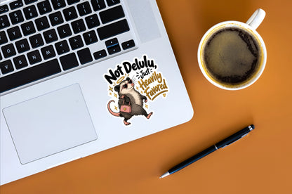 A die-cut sticker featuring a cartoon badger carrying a pink bag with the text 'Not Delulu, Just Heavily Favored' applied to the palm rest of a silver laptop. The laptop is shown in a styled flat-lay on an orange surface alongside a cup of coffee and a black pen.