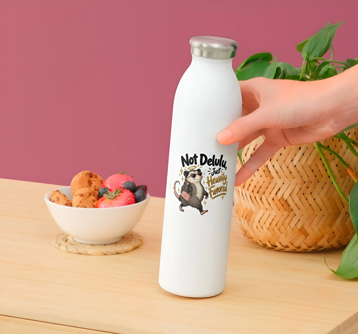 A hand holds a white insulated water bottle with a silver cap on a wooden table. The bottle has a cartoon illustration of a raccoon and the text 'Not Delays Just Heavy Drinking'.