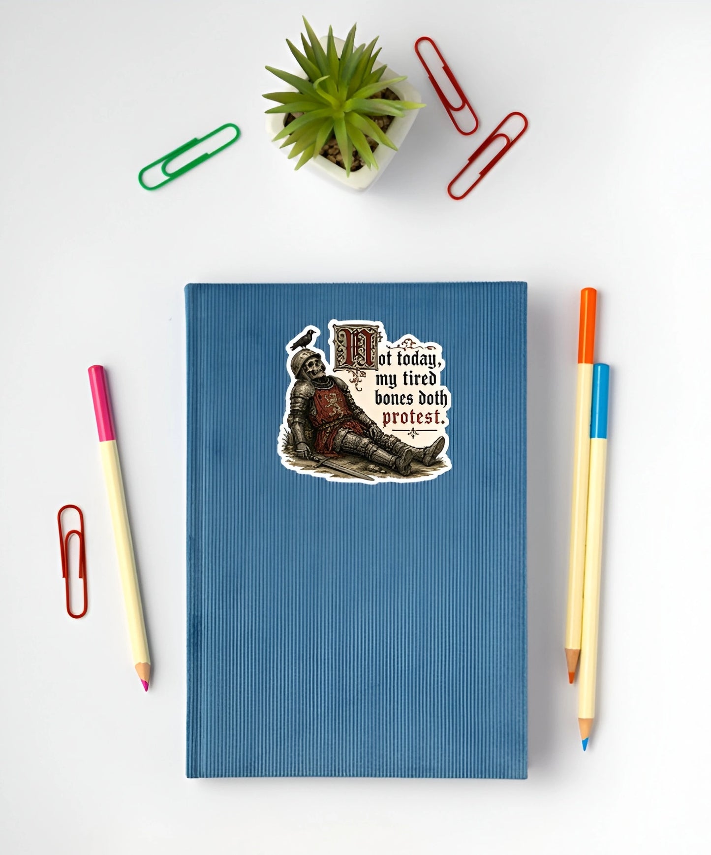 A blue ribbed notebook or journal with a die-cut sticker on the front, styled in a flat lay with a small potted succulent, paperclips, and colored pencils on a white background.