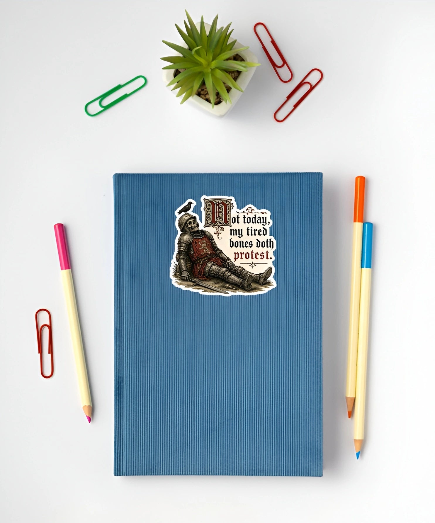 A blue ribbed notebook or journal with a die-cut sticker on the front, styled in a flat lay with a small potted succulent, paperclips, and colored pencils on a white background.