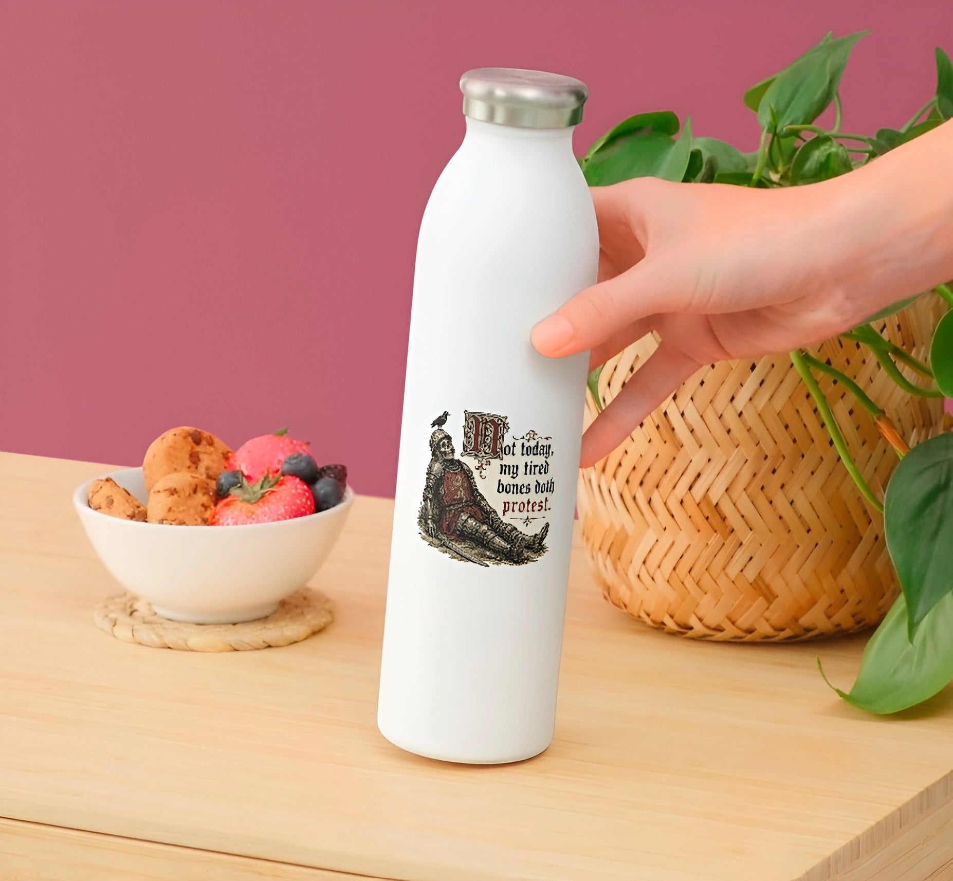 A hand holds a white insulated water bottle with a silver cap on a wooden table. The bottle has a black graphic of a knight skeleton